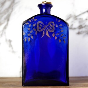 May include: A deep blue glass bottle with a rectangular form and a gold-coloured metal ring around the neck. The bottle is decorated with a gold bow and leaf design. The bottle is on a wooden surface.