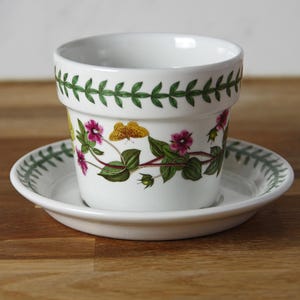 May include: White ceramic plant pot and saucer set. The pot features a floral and butterfly design in pink, yellow, and green. The rim of the pot and saucer have a green leaf pattern. A decorative item.