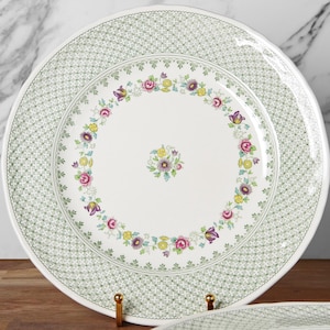 May include: White ceramic plates with a green geometric border and a floral design. The plates feature a central floral arrangement and a ring of flowers in pink, yellow, and purple. The plates are displayed on a wooden surface.