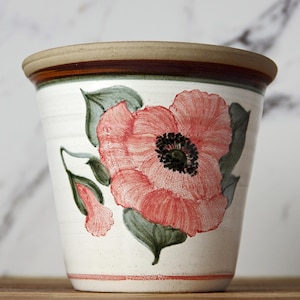 May include: A white ceramic plant pot featuring a hand-painted pink poppy flower with green leaves. The rim is tan and brown. Suitable for indoor or outdoor use. The pot's height is approximately 15 cm.