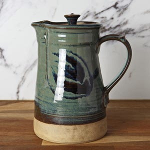 May include: A handcrafted ceramic jug with a lid. The jug is glazed in shades of teal, blue, and brown, with a cream-coloured base. It has a curved handle and a spout for pouring. The jug is likely used for serving beverages.