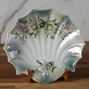 May include: A scalloped, white ceramic dish with a hand-painted floral design featuring daisies. The dish has a pearlescent finish and is supported by a gold-coloured stand. The edges are trimmed with a teal-coloured border.