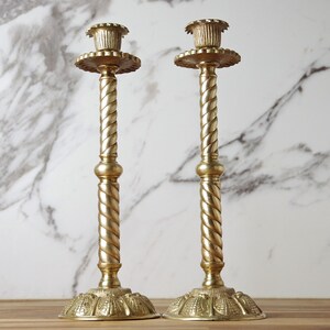 May include: A pair of tall, polished brass candlesticks featuring a twisted rope-style stem, a circular base with embossed detailing, and a fluted candle cup at the top. The metal has a warm, reflective gold-toned finish throughout the structure.