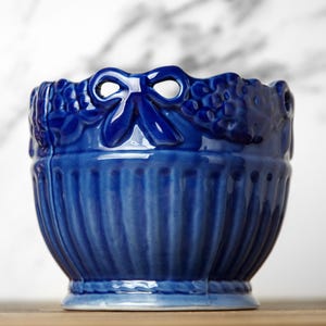 May include: A deep blue ceramic planter featuring a vertical ribbed texture on the lower body and a decorative rim adorned with sculpted bows and floral patterns. The vessel has a glossy finish and a wide, open top, showcasing a solid colour.
