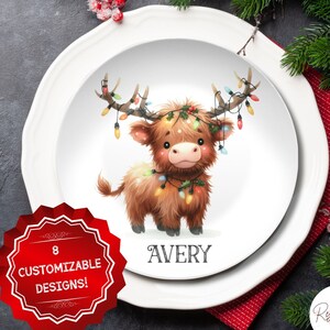 May include: White ceramic dinner plate featuring a cartoon illustration of a brown Highland cow with Christmas lights wrapped around its antlers and neck. The plate has the name "AVERY" printed in black below the illustration.