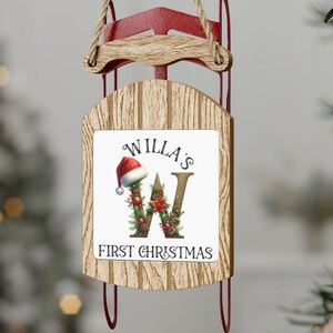 May include: A wooden sleigh Christmas ornament with a white square on the front. The square has the text "Willa's First Christmas" and a decorated letter "W" with a Santa hat.