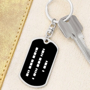 May include: A black and silver keychain with a dog tag featuring a white braille pattern. The keychain has a silver ring and a silver key attached.
