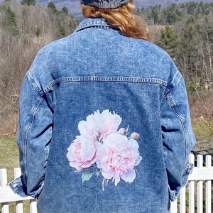 A person wearing a denim jacket with a floral design on the back, standing in front of a white picket fence and a mountainous landscape.
