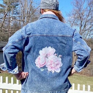 A person wearing a denim jacket with a floral design on the back stands in front of a white picket fence and trees.