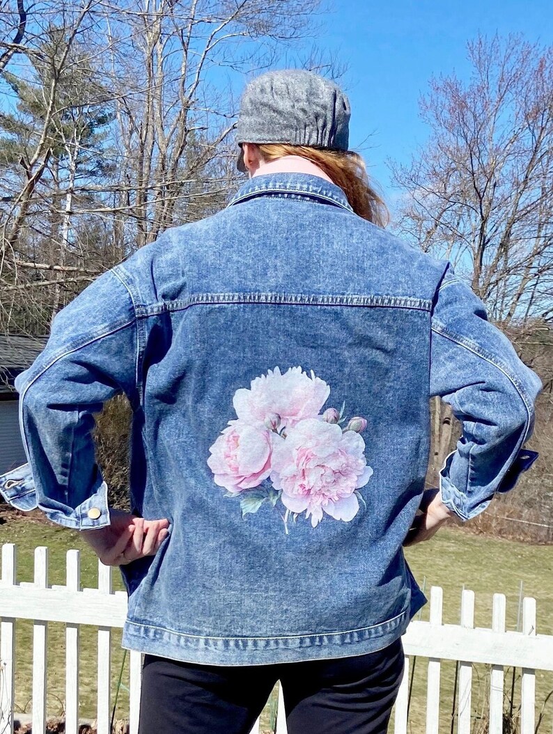 A person wearing a denim jacket with a floral design on the back stands in front of a white picket fence and trees.