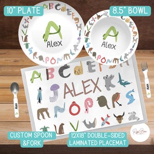 May include: A personalized children's placemat set with a 10-inch plate, 8.5-inch bowl, spoon, and fork. The placemat features a white background with colorful letters, animals, and the name "Alex".