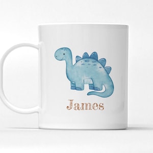 Peut inclure: Mug en c&eacute;ramique blanche avec une illustration de dinosaure bleu clair &agrave; l'aquarelle. Le dinosaure a une expression amicale et est &eacute;tiquet&eacute; avec le nom "James" dans une police d&eacute;corative marron. La tasse a une anse incurv&eacute;e.