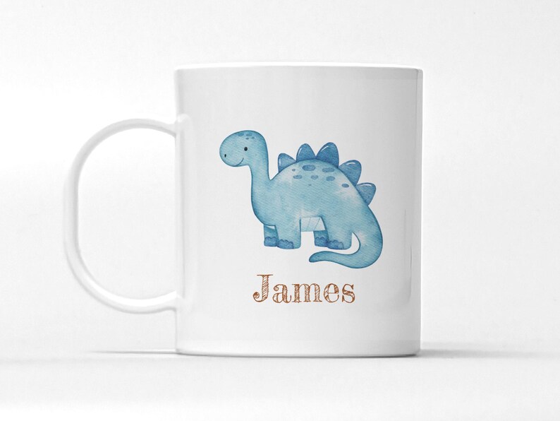 Peut inclure: Mug en c&eacute;ramique blanche avec une illustration de dinosaure bleu clair &agrave; l'aquarelle. Le dinosaure a une expression amicale et est &eacute;tiquet&eacute; avec le nom "James" dans une police d&eacute;corative marron. La tasse a une anse incurv&eacute;e.