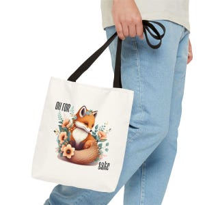 A person is holding a white tote bag with a printed design of a fox surrounded by flowers and leaves.