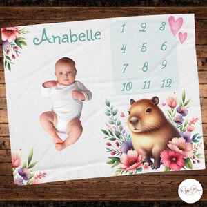 May include: White baby milestone blanket with the name "Anabelle" in teal script. Features a baby, a capybara illustration surrounded by flowers, and a monthly tracker with numbers 1-12. Includes pink hearts.