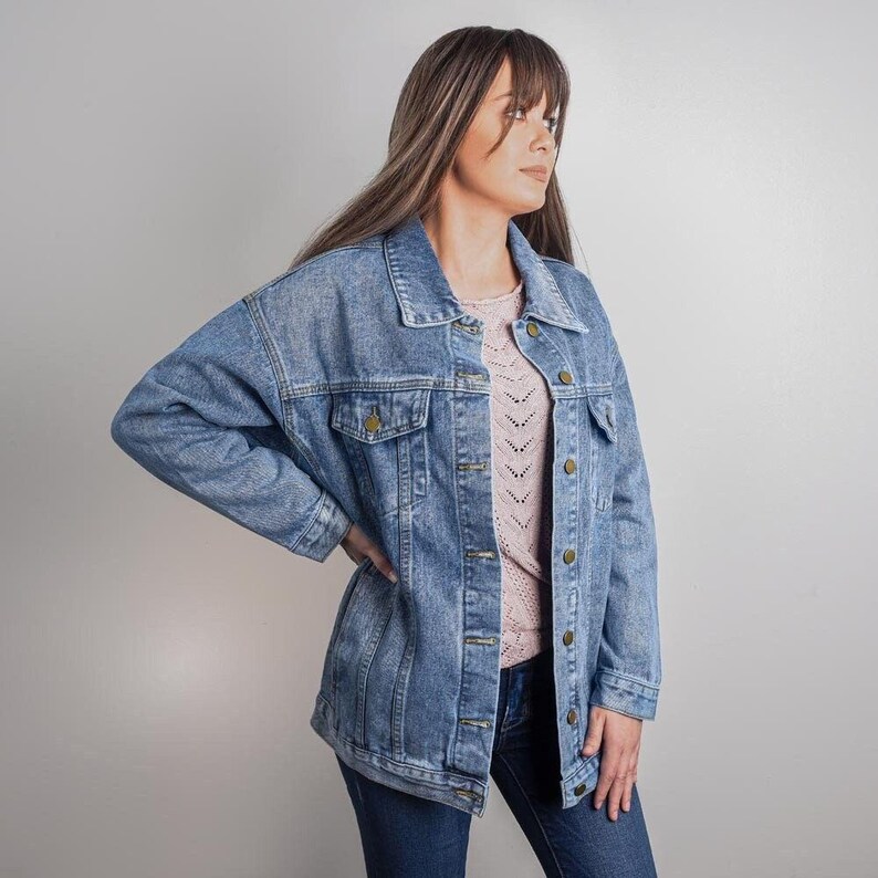 A woman wearing a blue denim jacket and a pink sweater stands against a plain background.