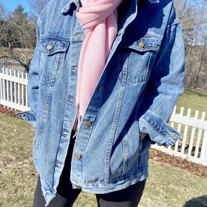 A person wearing a denim jacket and a pink scarf stands in front of a white picket fence, with trees and a blue sky in the background.