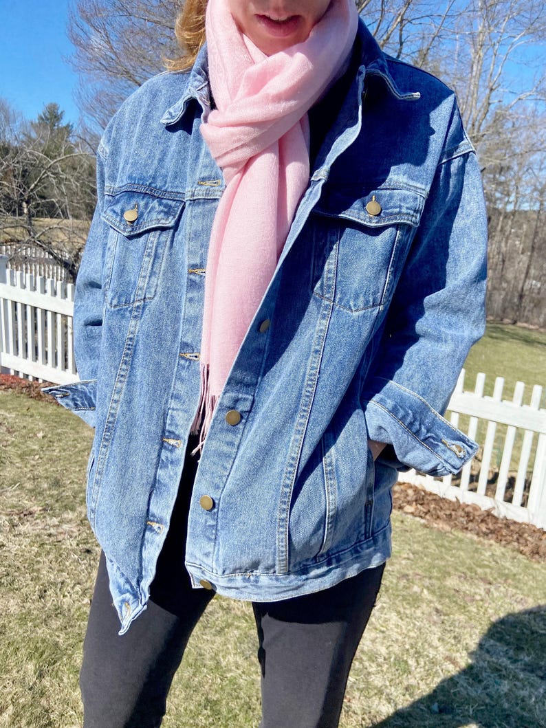 A person wearing a denim jacket and a pink scarf stands in front of a white picket fence, with trees and a blue sky in the background.