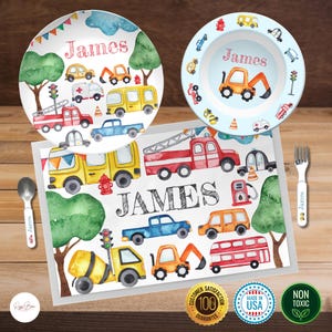 May include: A personalized children's place setting with a watercolor design featuring a variety of vehicles, including a fire truck, bus, ambulance, and construction vehicles. The place setting includes a plate, bowl, and placemat, all with the name "James" printed on them.