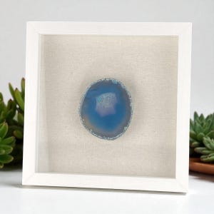 May include: A framed blue agate slice. The polished stone is set against a neutral fabric background within a white square frame. The agate features shades of blue and a crystalline center, creating a decorative piece.
