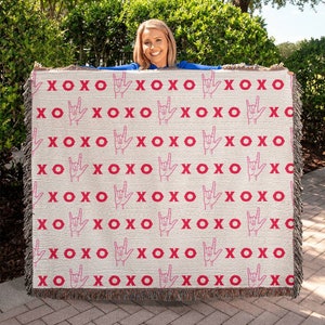 May include: A woven throw blanket with a white background and a repeating pattern of pink XOXO and pink hand signs for 'I love you' in American Sign Language.