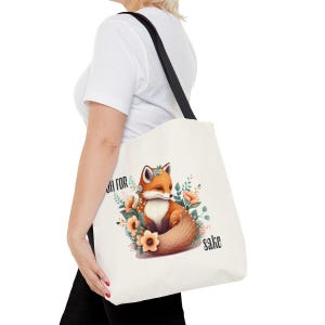 A person is holding a white tote bag with a printed design of a fox surrounded by flowers and leaves.