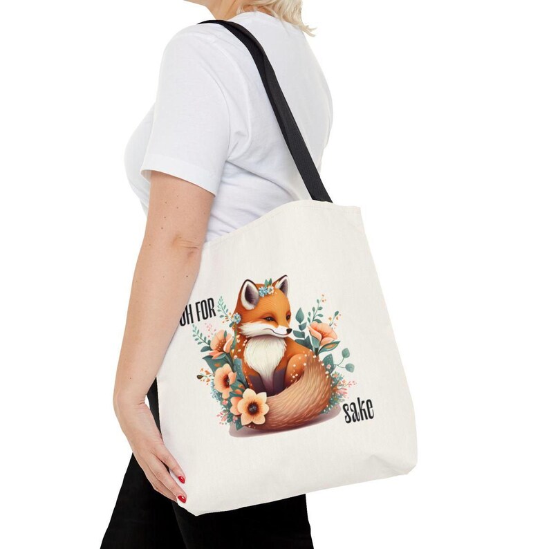 A person is holding a white tote bag with a printed design of a fox surrounded by flowers and leaves.
