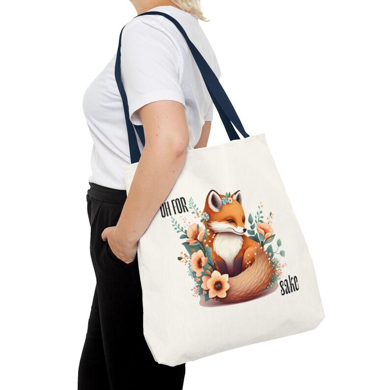A person is holding a white tote bag with a printed design of a fox surrounded by flowers.