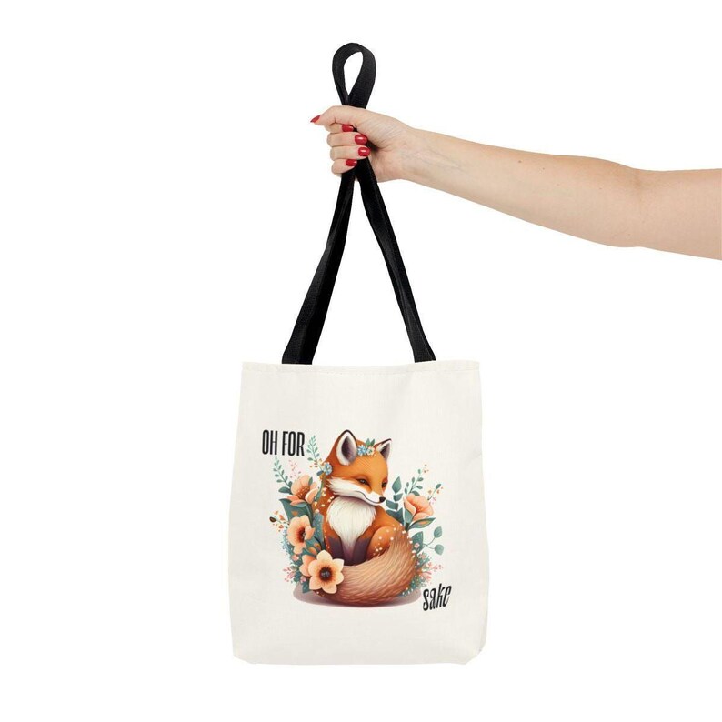 A hand holding a white tote bag with a printed image of a fox surrounded by flowers.