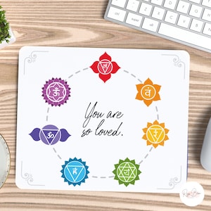 May include: White mouse pad with a circle of seven colorful chakras, each with a Sanskrit symbol. The text "You are so loved." is written in black cursive script in the center of the circle.