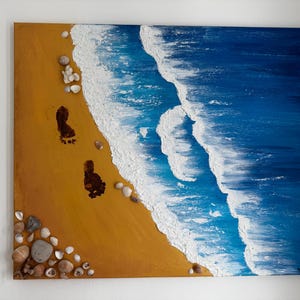 Könnte beinhalten: Ein Strandgemälde mit goldenem Sand, braunen Fußabdrücken und Muscheln. Das Meer ist in Blautönen mit weißen Wellen dargestellt. Das Kunstwerk ist eine quadratische Leinwand.