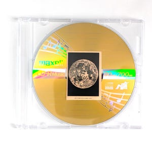 Puede incluir: Un disco CD-R dorado con una pequeña tarjeta con una ilustración detallada de la luna. El disco está en una funda de plástico transparente. El disco tiene el texto "maxell" y "CD-R Music" impreso.