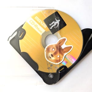 May include: A gold CD-R disc with a bunny sticker and a black case. The disc is labeled "maxell CD-R Music". A small sticker of a person is on the disc. The case is clear plastic.