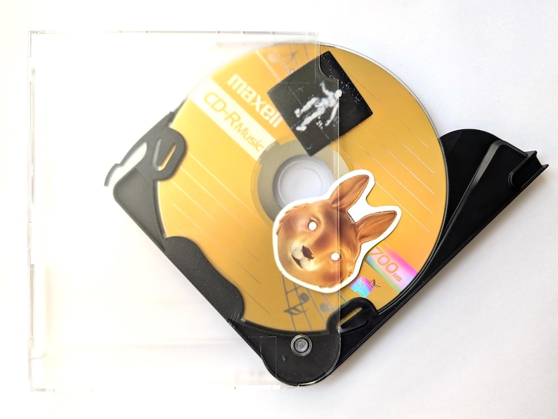 May include: A gold CD-R disc with a bunny sticker and a black case. The disc is labeled "maxell CD-R Music". A small sticker of a person is on the disc. The case is clear plastic.