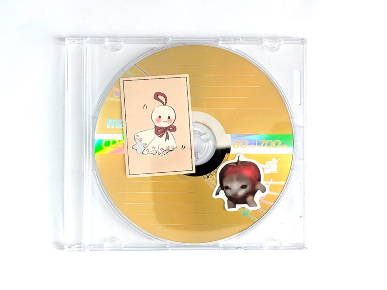 May include: A gold CD in a clear plastic case with two stickers. One sticker is a tan rectangle with a cartoon doll illustration. The other sticker is a cartoon apple with a cat face. The CD has the text "80-1700".