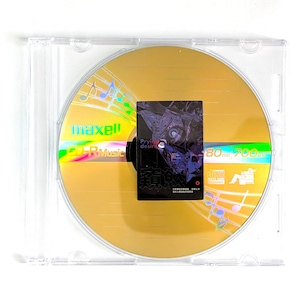May include: A gold-colored CD-R disc with a holographic rainbow sheen, encased in a clear plastic jewel case. The disc has the brand name "maxell" and "CD-R Music" printed on it. A small black square with text and an illustration is affixed to the disc.