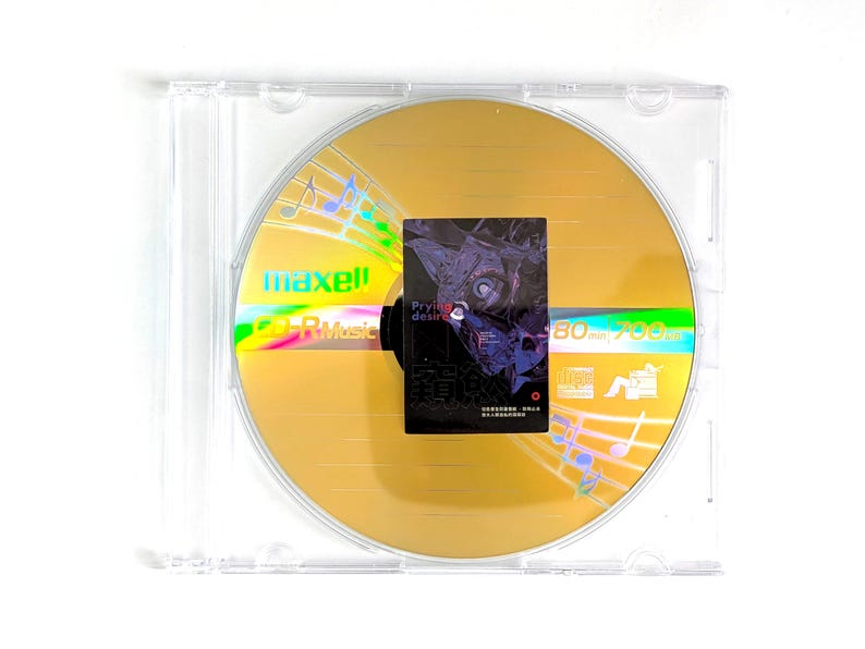 May include: A gold-colored CD-R disc with a holographic rainbow sheen, encased in a clear plastic jewel case. The disc has the brand name "maxell" and "CD-R Music" printed on it. A small black square with text and an illustration is affixed to the disc.