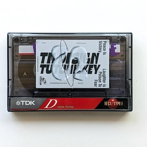 Peut inclure: Une cassette transparente avec une étiquette rouge et noire indiquant "TDK D". La cassette a une étiquette blanche avec le texte "TEN TUKEY 82 DESIGN". L'étiquette comprend également les phrases "Peace Is Victory" et "Laughter Is Poison To Fear".