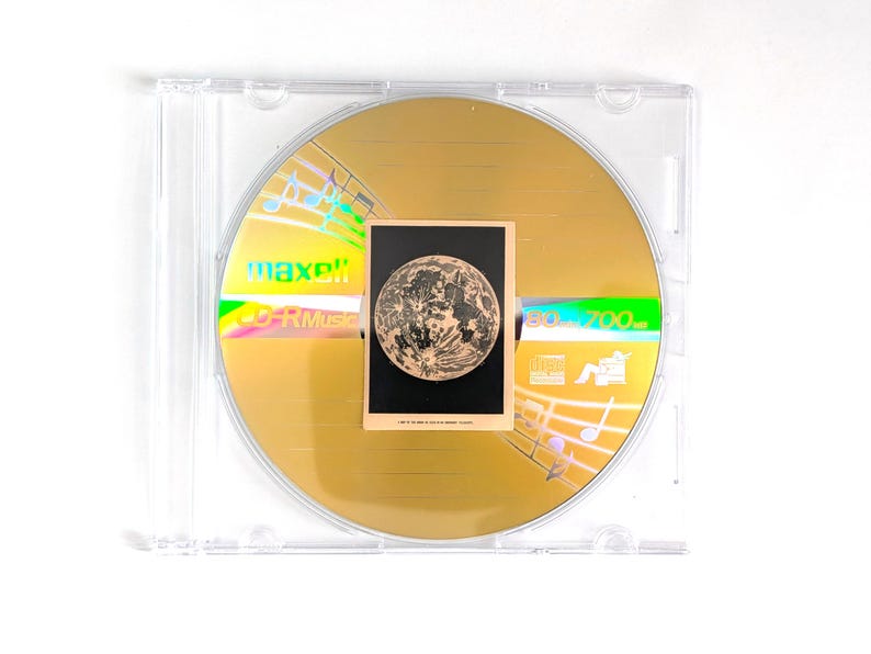May include: A gold-colored CD-R disc with a small card featuring a detailed illustration of the moon. The disc is in a clear plastic case. The disc has the text "maxell" and "CD-R Music" printed on it.
