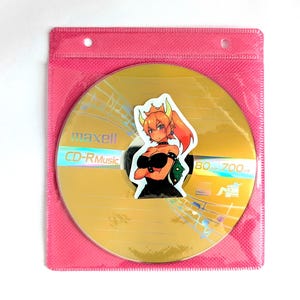 May include: A gold Maxell CD-R Music disc with a sticker of an anime character in a pink plastic sleeve. The disc has musical notes and the text "CD-R Music" printed on it. The sticker features a female character with orange hair and a black outfit.