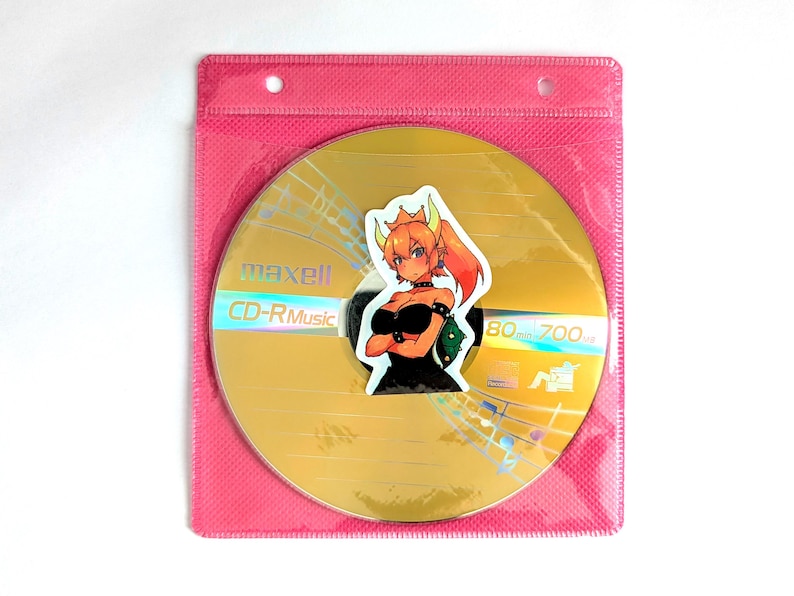 May include: A gold Maxell CD-R Music disc with a sticker of an anime character in a pink plastic sleeve. The disc has musical notes and the text "CD-R Music" printed on it. The sticker features a female character with orange hair and a black outfit.