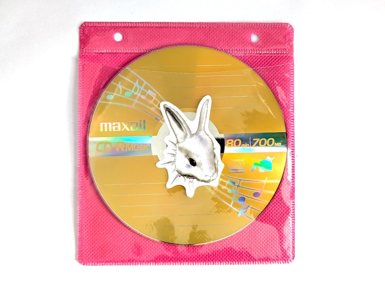May include: A gold CD-R disc with a rabbit sticker, inside a pink plastic sleeve. The disc has musical notes and the text "maxell CD-R Music". The sticker is a white rabbit head. The disc is 80 min / 700 MB.
