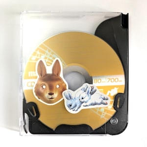 May include: A CD in a clear plastic case. The CD is gold with musical notes and two stickers: a brown rabbit head and two blue and white rabbits. The CD has the text "80 mm 700 MB".