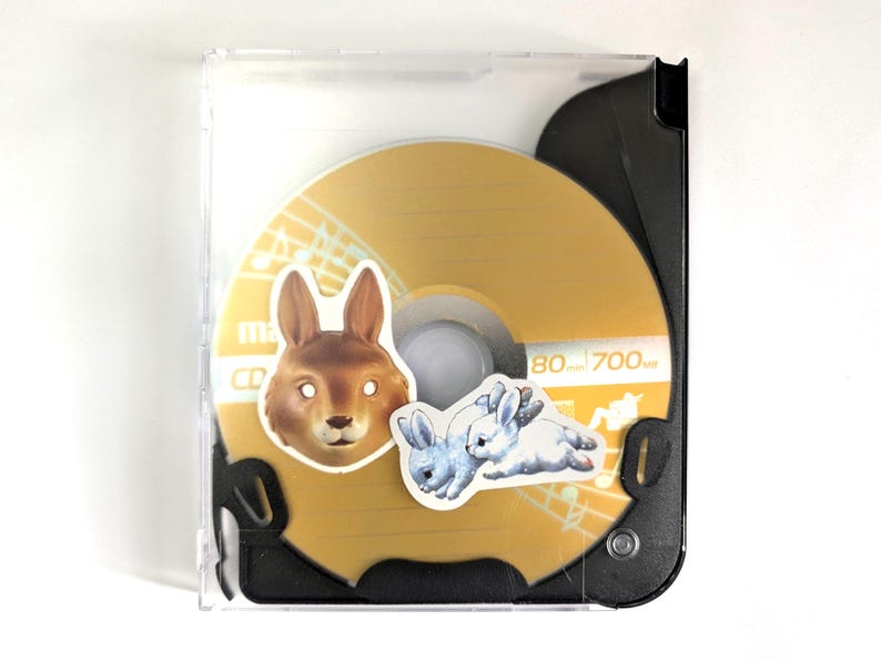 May include: A CD in a clear plastic case. The CD is gold with musical notes and two stickers: a brown rabbit head and two blue and white rabbits. The CD has the text "80 mm 700 MB".