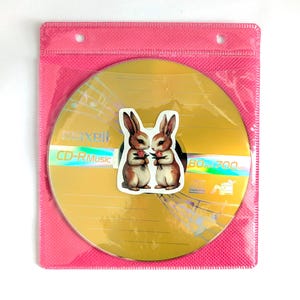 May include: A gold CD-R disc with the text "CD-R Music" and a sticker of two brown and white bunnies holding flowers. The disc is in a pink plastic sleeve.