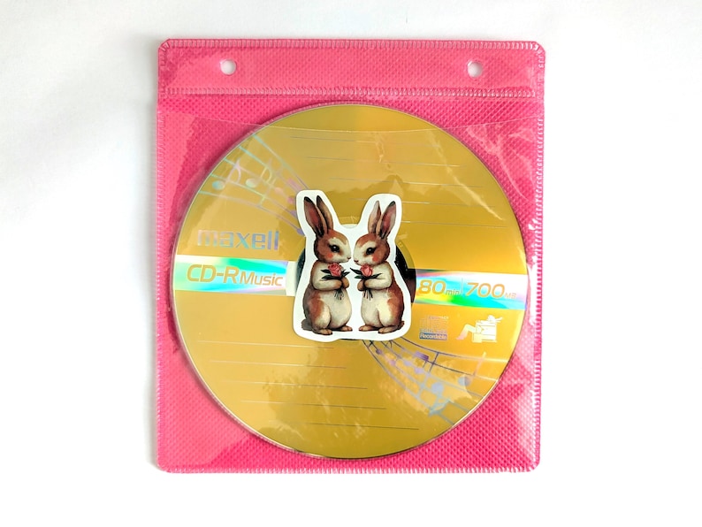 May include: A gold CD-R disc with the text "CD-R Music" and a sticker of two brown and white bunnies holding flowers. The disc is in a pink plastic sleeve.