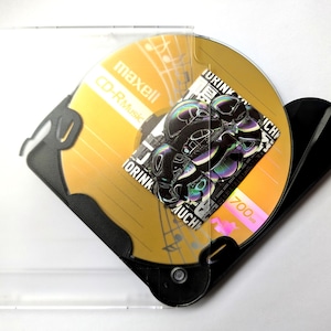 May include: A gold Maxell CD-R Music disc with a black and white graphic design, inside a clear plastic case. The disc has the text "TORINK" and "700MB" printed on it. The case is partially open.