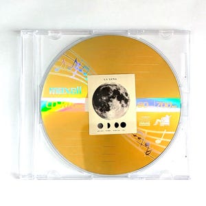 May include: A gold-colored CD in a clear plastic case. The CD has the word "maxell" printed on it. A small card with a moon graphic and the words "LA LUNA" is on top of the CD. Musical notes are printed on the CD.