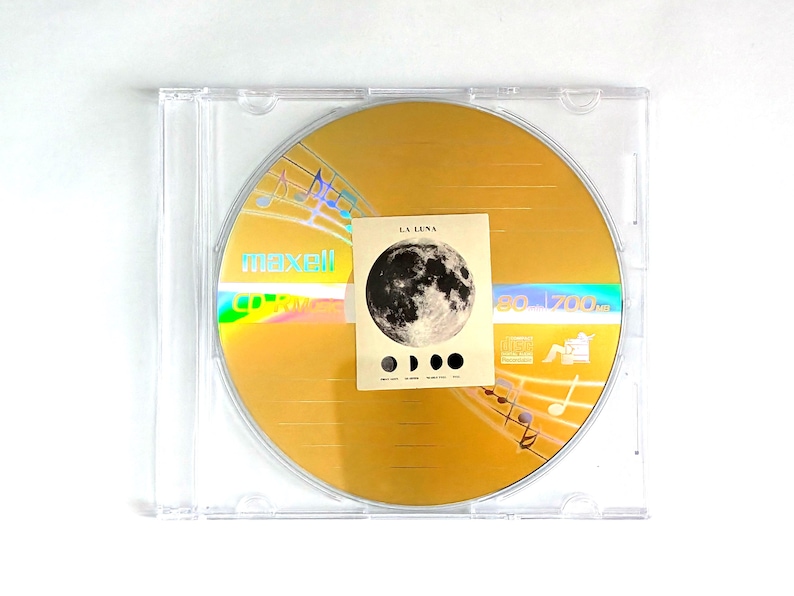 May include: A gold-colored CD in a clear plastic case. The CD has the word "maxell" printed on it. A small card with a moon graphic and the words "LA LUNA" is on top of the CD. Musical notes are printed on the CD.