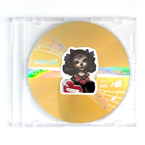 May include: A Maxell CD-R disc in a clear plastic case. The gold disc has a sticker of a black dog with a red heart collar and a heart-shaped sign that says "Hello!". The disc has musical notes printed on it.
